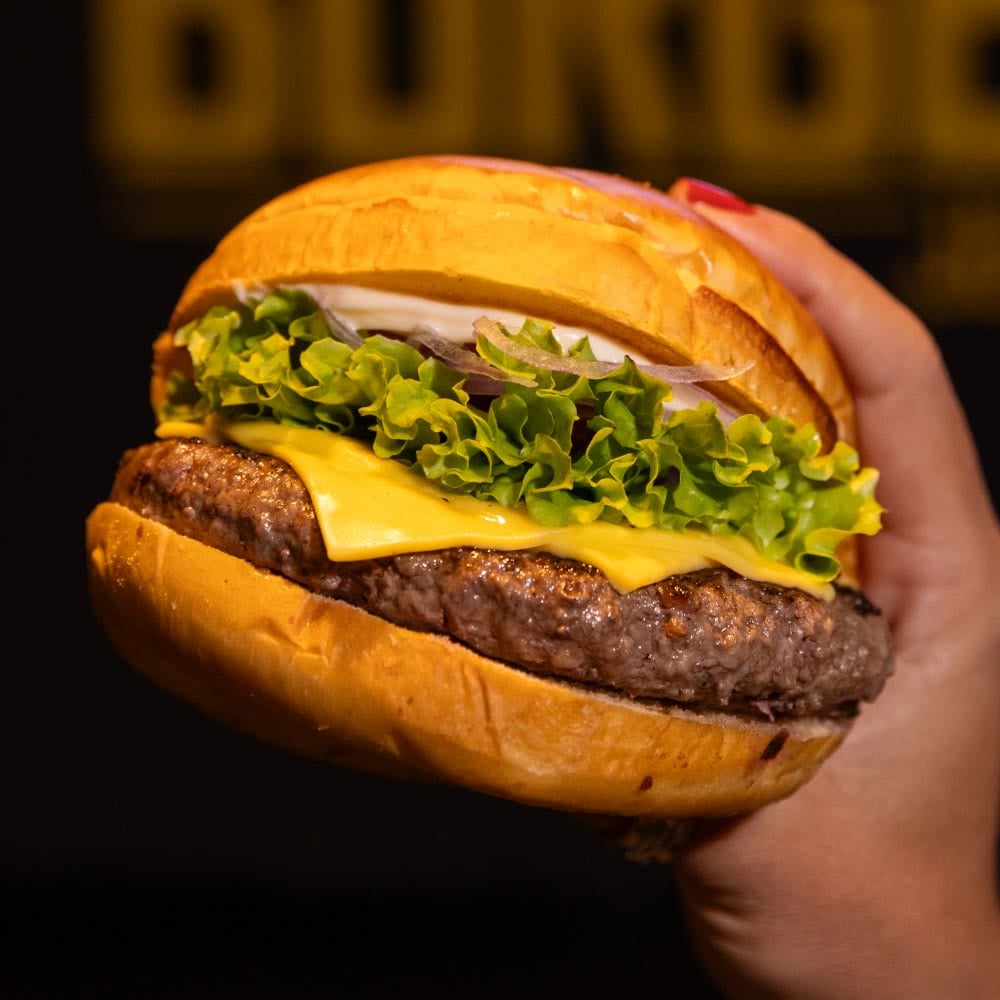Burger Jurerê na mão, com alface, queijo prato e cebola roxa, em frente ao letreiro do BBJ
